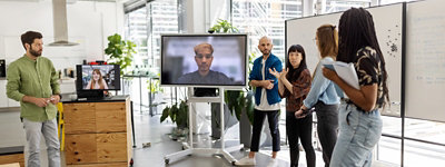 A team huddle in an office space with five people attending in person and two people dialing in from other locations via Teams.