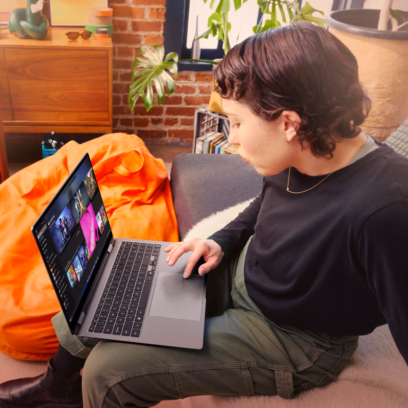 A person sitting on a sofa looking at PC games on an open laptop with a bean bag, amp, shelf and potted plant in the background.