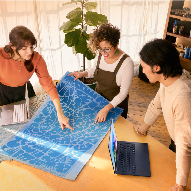 Three people, in an open, bright room with sheer curtained windows, plants and a bookshelf in the background, are leaning over a table with two open laptops to look at a large blue abstract print.